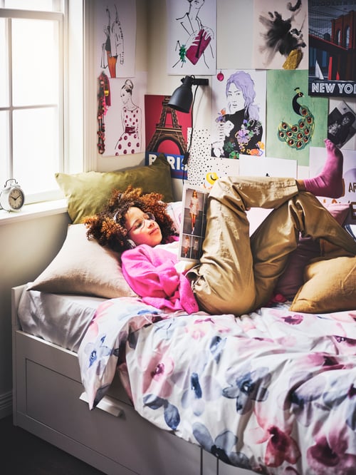 Kinderzimmer mit einem BRIMNES Tagesbettgestell, einem Kissen in einem AINA Kissenbezug und einem HEKTAR Wand-/Klemmspot. Auf dem Bett liegt ein Kind und liest.