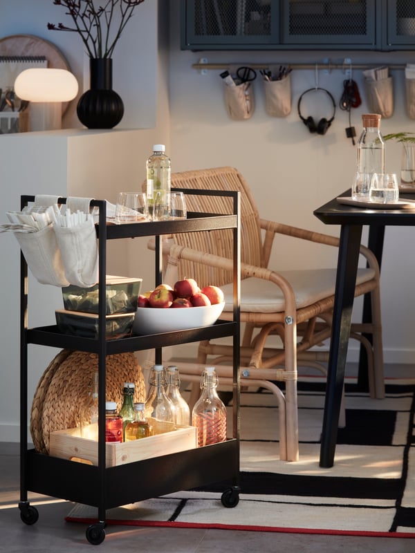 A softly sunlit dining area with a black NISSAFORS cart laden with dining accessories placed next to a black LISABO table.