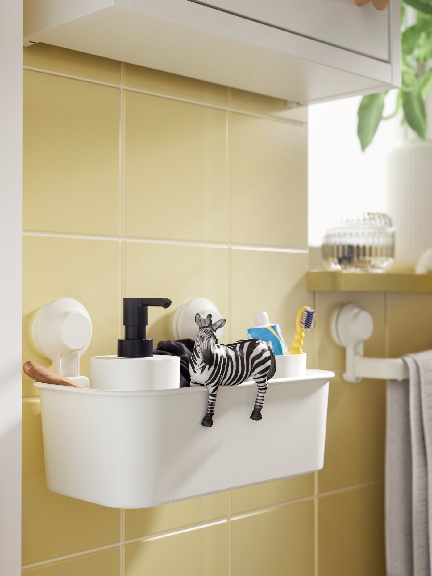 Badrum med vit förvaringslåda fäst på en gulkaklad vägg. I lådan finns bland annat en tvålpump, en zebraformad figur och tandborstar. Till höger hänger en handduk.