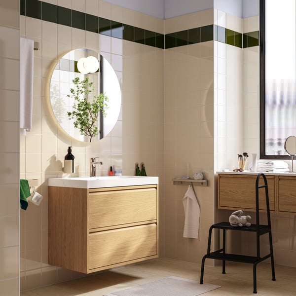 An oak effect ÄNGSJÖN/BACKSJÖN wash-stand with two drawers and a wash-basin is below a circular mirror in a modern bathroom.