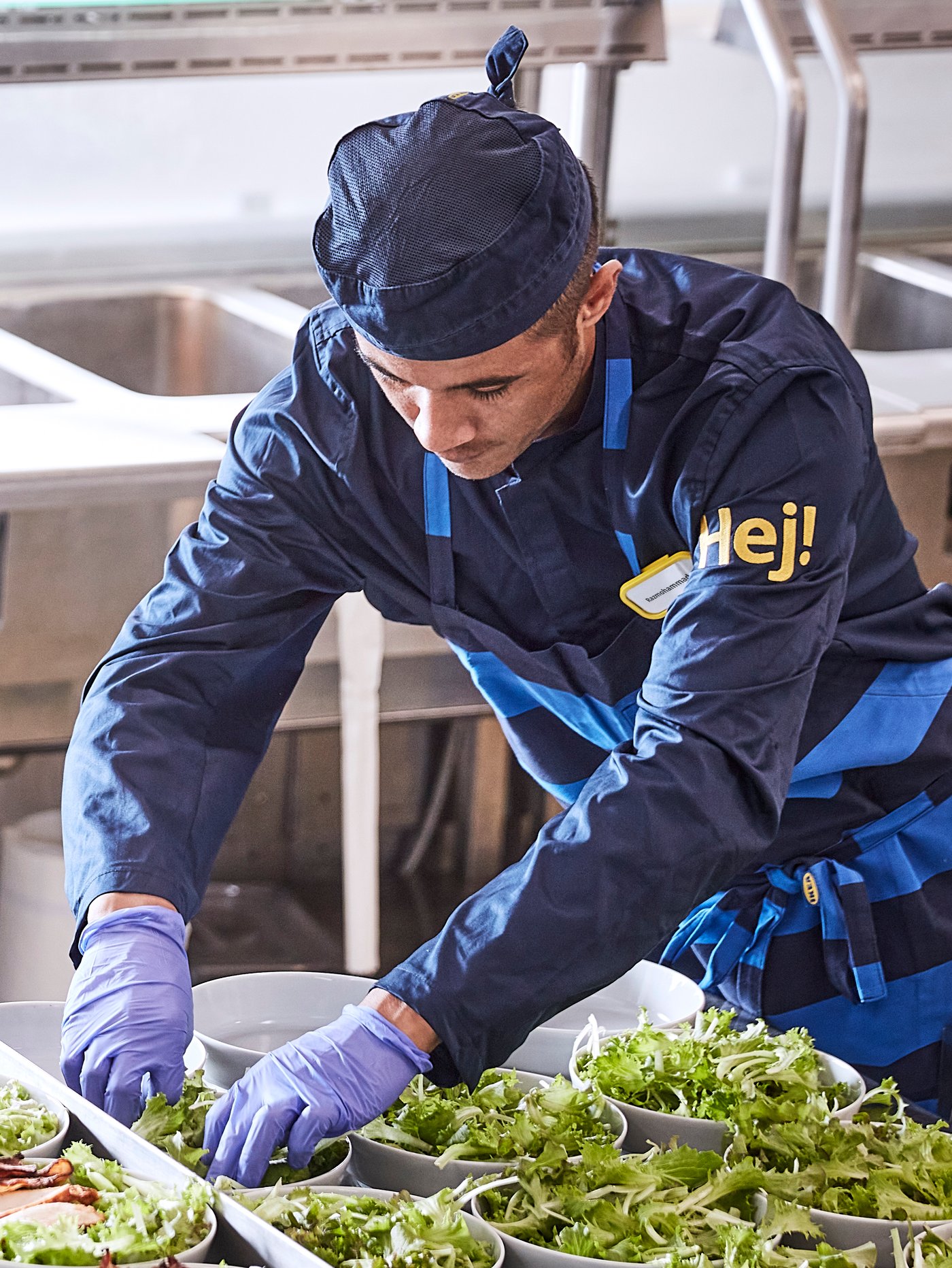 A person in a restaurant kitchen wearing work clothes, gloves and an apron is filling bowls in a row with salad.