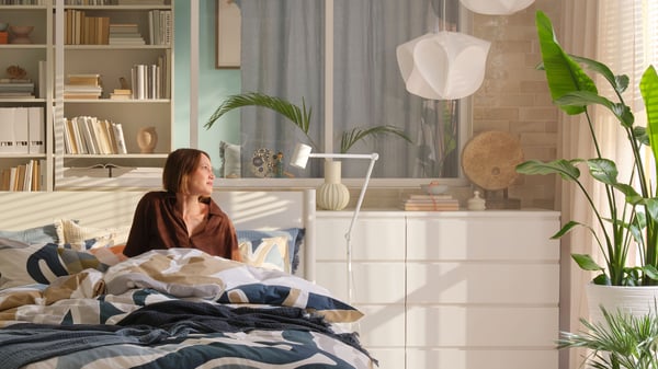 A bright bedroom with white MALM chests of drawers and BILLY bookshelves, a woman sits in a TONSTAD bed gazing out through the window.