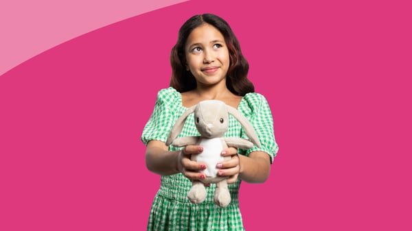 Bambina in abito verde a quadretti che porge un peluche a forma di coniglio su sfondo rosa acceso.