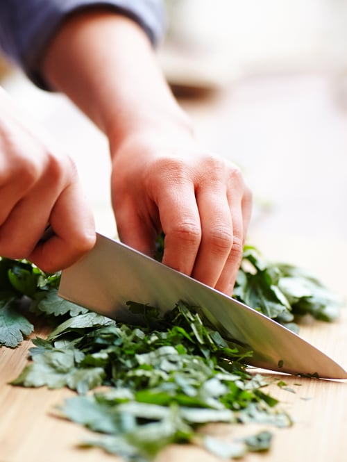 A pair of hands cutting fresh herbs with a kitchen knife on top of a wood cutting board.