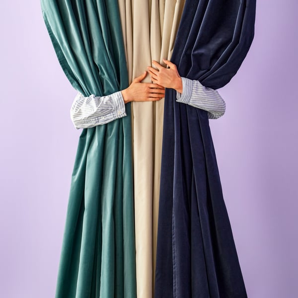 A person embracing three SANELA curtains from behind. The curtains are dark blue, dark grey-turquoise and light beige.