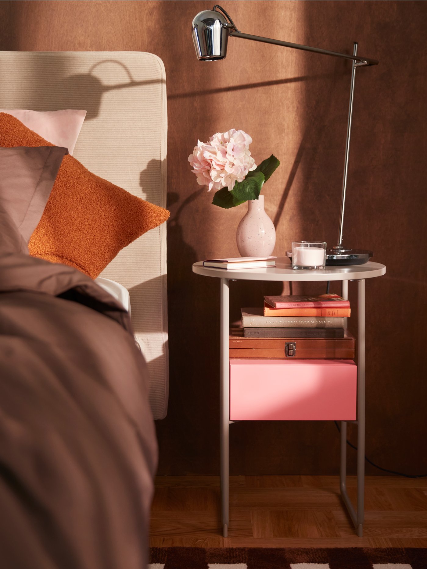 A bedroom with a beige/pink BLANKHÄLLAN bedside table beside a bed, topped with flowers and a lamp against a brown wall.