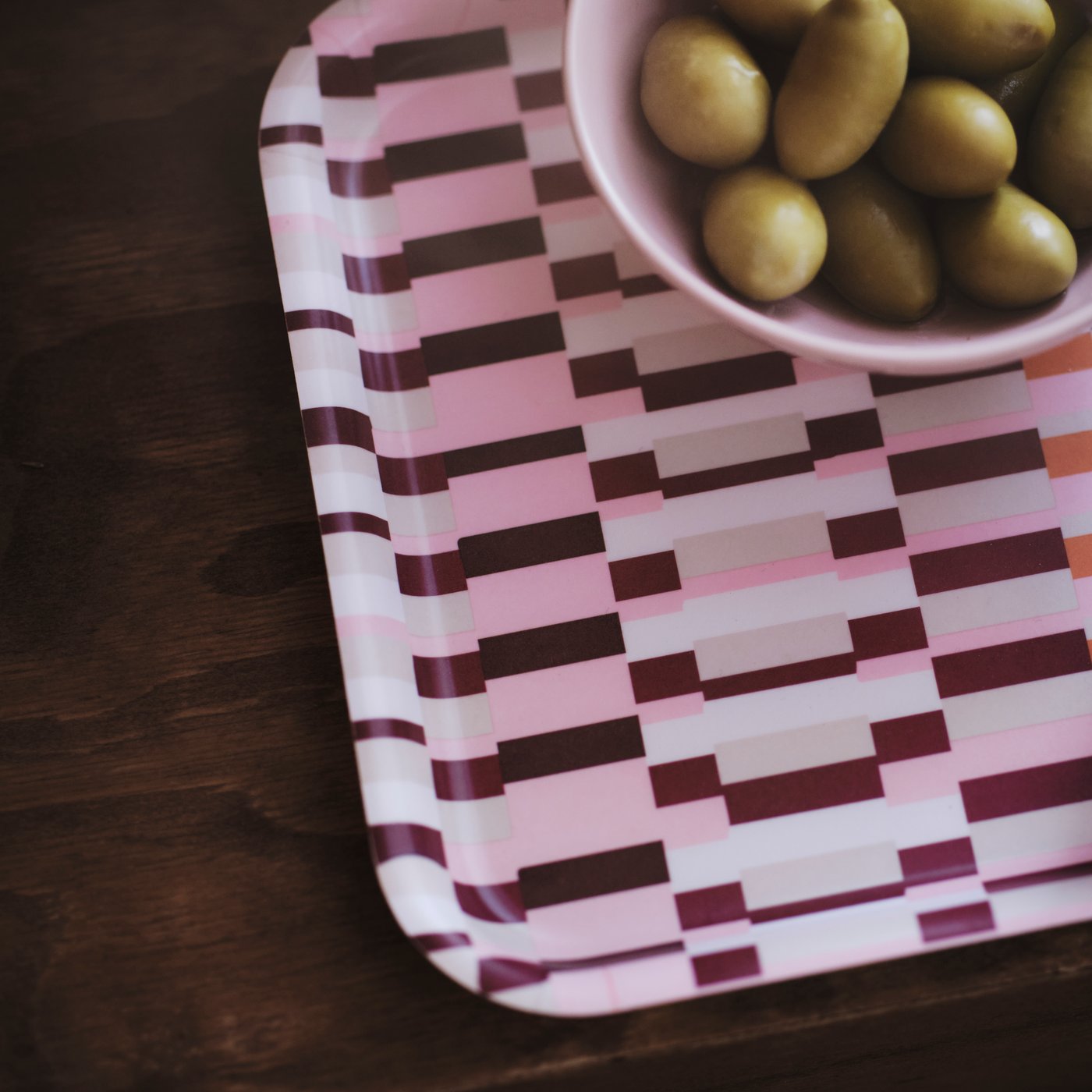 A close-up of a pink patterned HÖSTAGILLE tray with a bowl of green olives on a dark wood table.