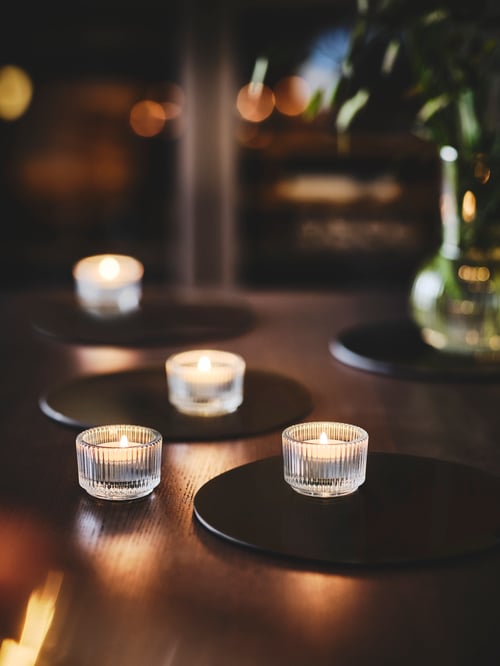 Sufragerie cu mai multe suporturi pentru tealight FINSMAK din sticlă transparentă albă care răspândesc o lumină caldă pe o masă din lemn.