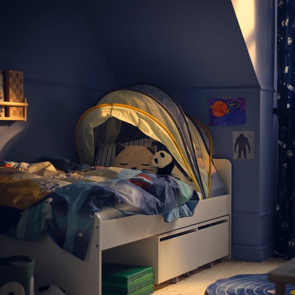 The corner of a childs bedroom with blue walls, a white SLÄKT bed with ELDFLUGA bed tent.