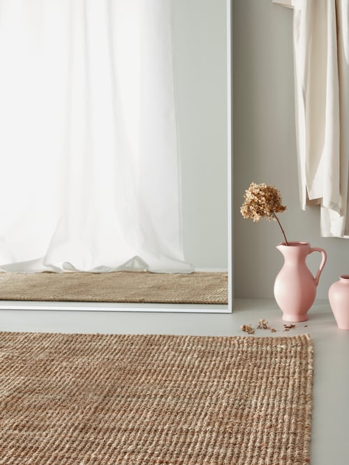Living room with a flatwoven rug on the floor, a pink vase with a single-stem flower, and a mirror that reflects curtains.