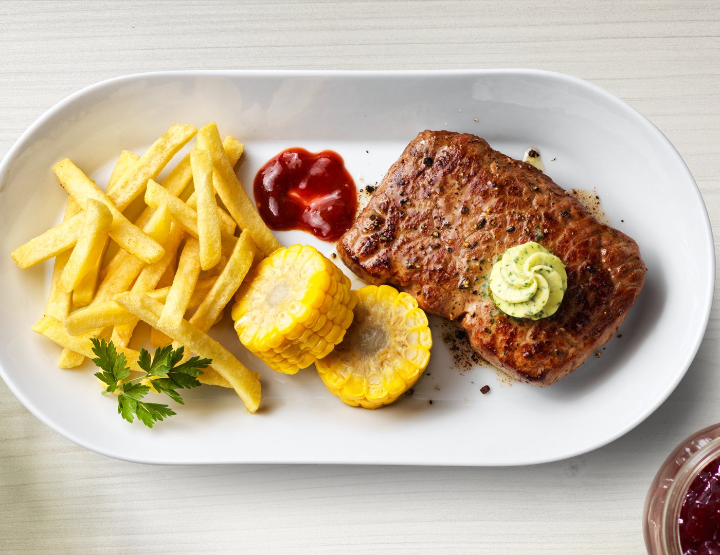 Auf einem weißen Teller sind ein gegrilltes Rumpsteak, Pommes frites und Maiskolbenscheiben angerichtet.