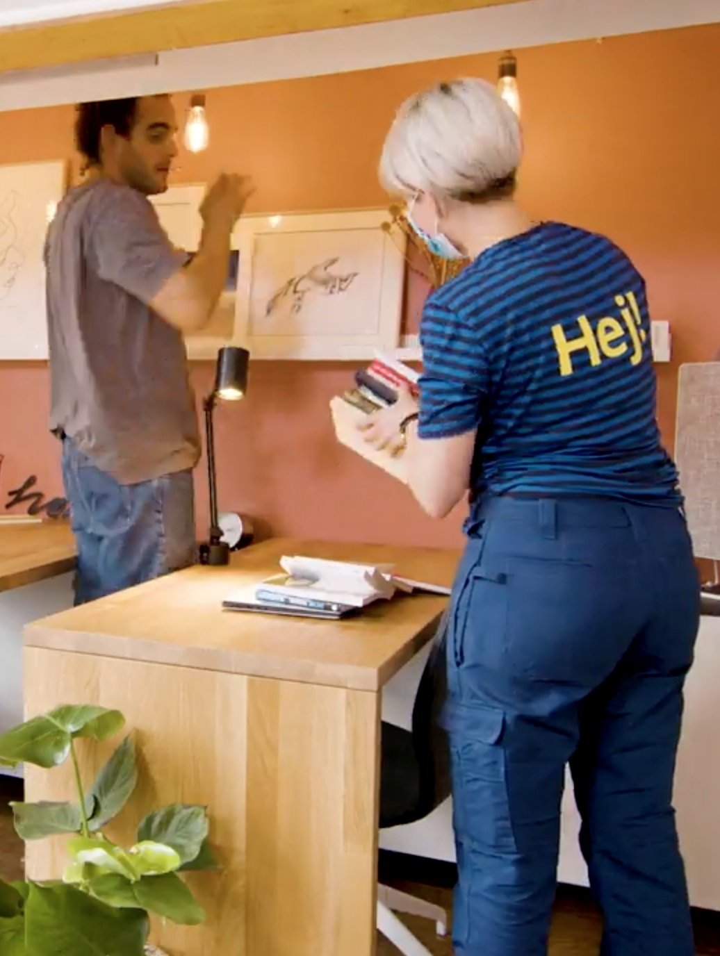  Muškarac stoji pred drvenim radnim stolom, na koji žena u IKEA uniformi stavlja gomilu knjiga i ram za slike.