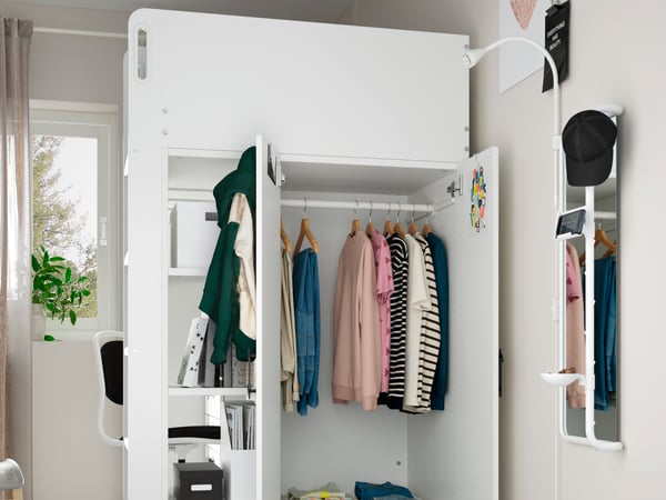 Organizador SMÅSTAD de color blanco con tres puertas, ropa de diferentes colores en su interior, cajas organizadoras blancas. Espejo y plantas en la parte trasera de la imagen.