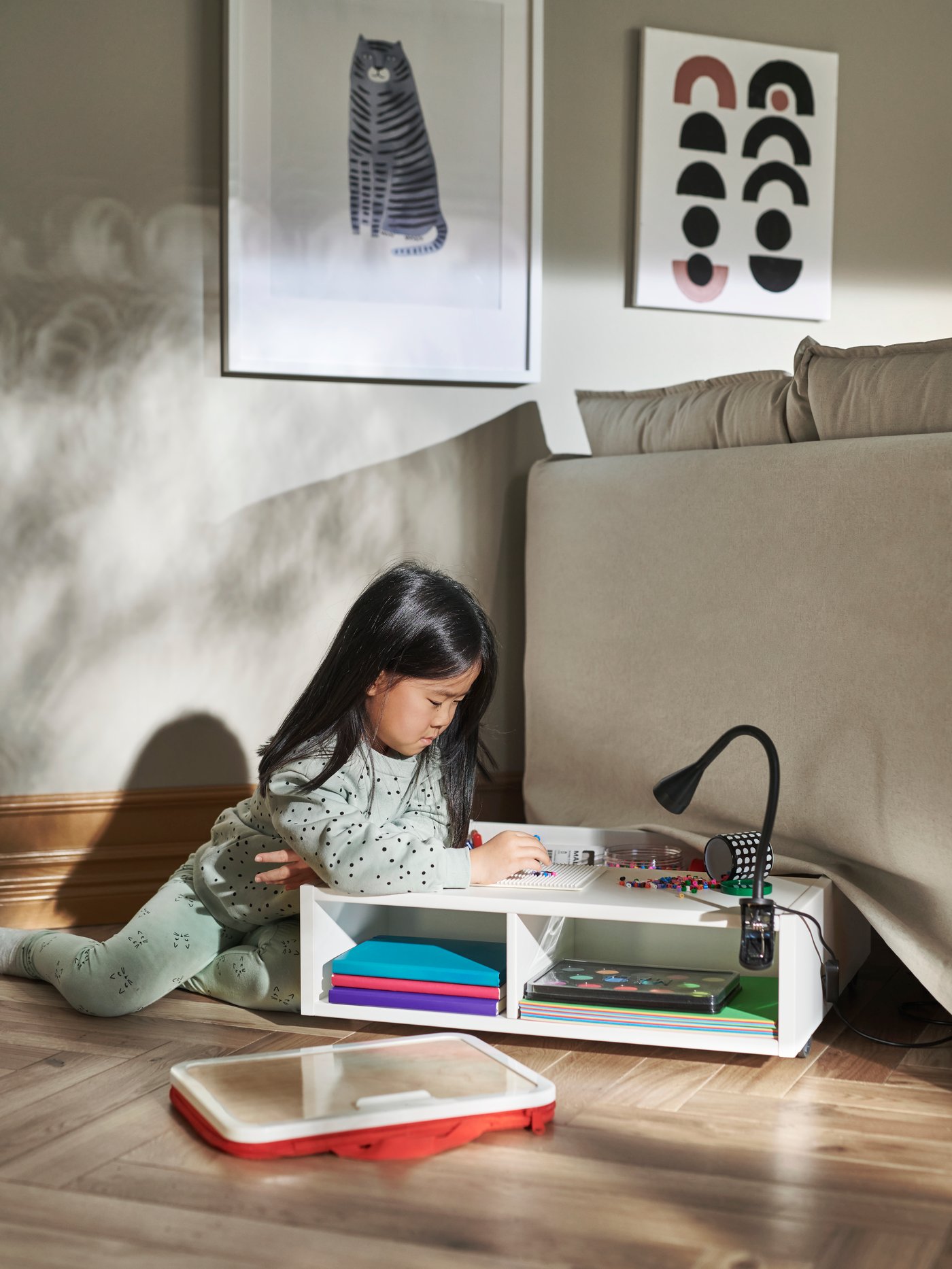 Una niña concentrada, apoyada sobre un cajón con ruedas FREDVANG blanco con artículos para hacer manualidades MÅLA. Sobre el cajón hay una lámpara de trabajo NÄVLINGE negra. 