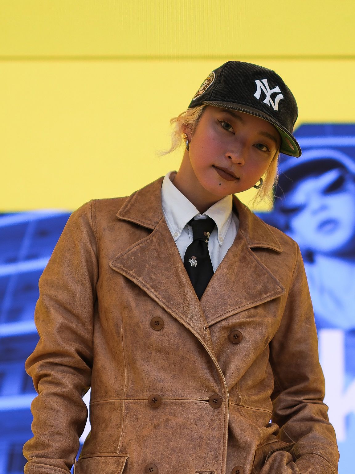 Kyoka, ki nosi usnjen plašč, kapo, kravato in belo srajco, stoji na odru pred ozadjem s svojo fotografijo v modri in rumeni barvi, za ozadje katero poskrbi digitalni zaslon, ki se med milanskim tednom oblikovanja 2024 uporablja na odru prizorišča IKEA. 