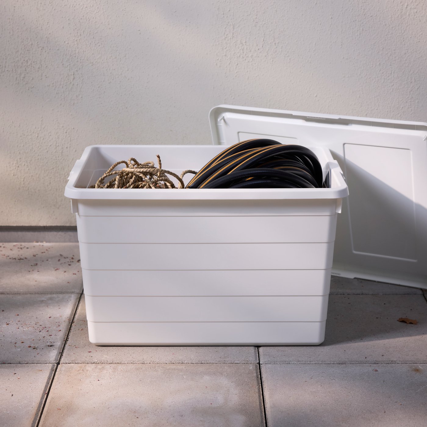 A white SOCKERBIT box with lid containing some rope and a garden hose stands on a terrace on a sunny day.
