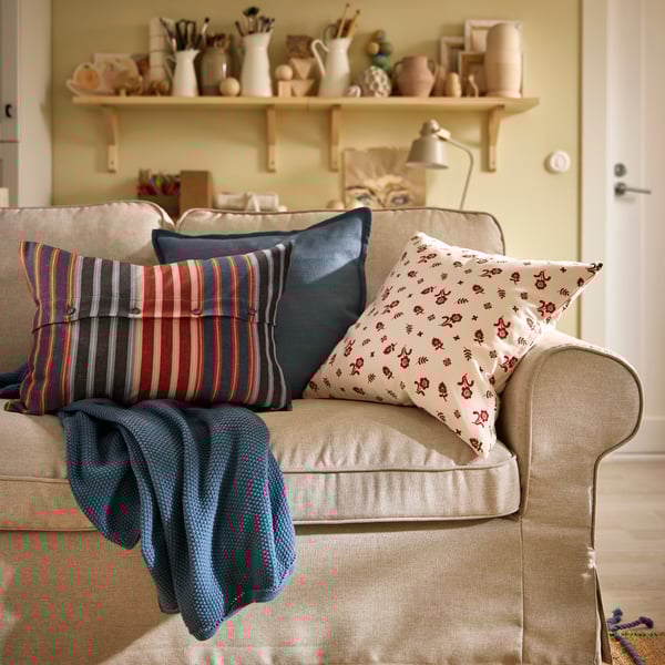 A blue-grey HUMLEMOTT throw and some cushions, including one with a floral TÅGSÄCKMAL cushion cover, lie on an EKTORP sofa.