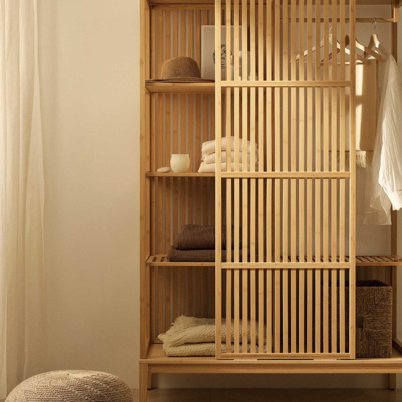 A beautifully organised NORDKISA open wardrobe with slatted sliding doors, perfect for keeping your belongings tidy and accessible. Inside, you'll find neatly folded textiles in warm, natural tones, a stylish woven hat, and a crisp white shirt hanging, showing off the versatile storage options. A cosy woven pouf sits on the wooden floor next to the wardrobe, offering a comfortable spot to sit or rest. This piece brings a touch of natural charm and practical elegance to any room.
