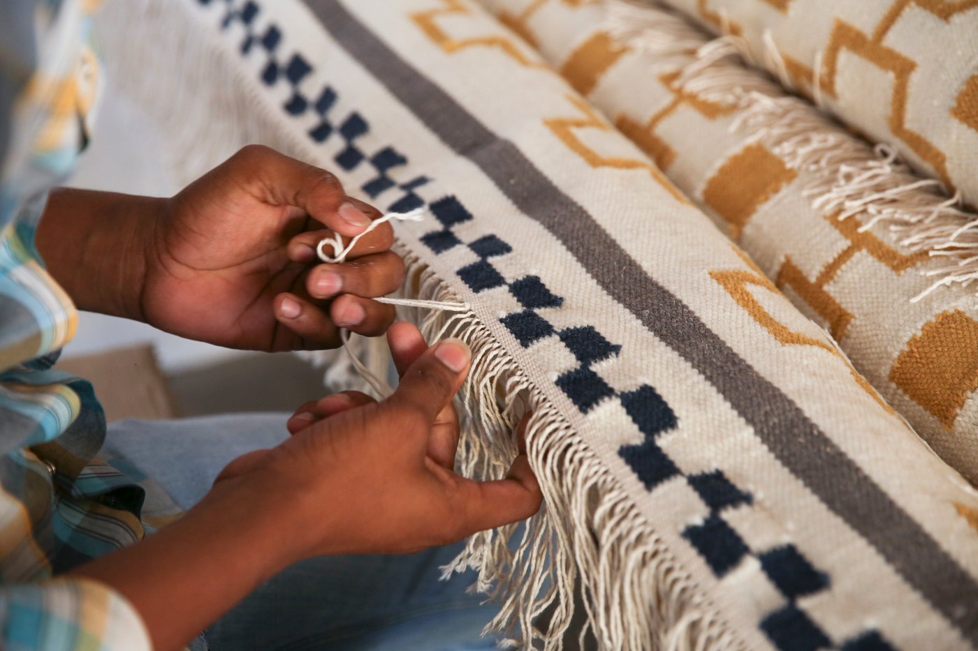 The hands of an Indian artisan weaving the fringe of a rug in shade of white, blue, grey and mustard.