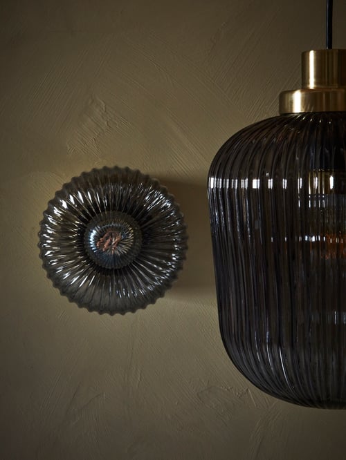 Lovitură parțială a unei lămpi cu pandantiv gri cu design din sticlă striată și o lampă montată pe perete cu design cu nervuri.