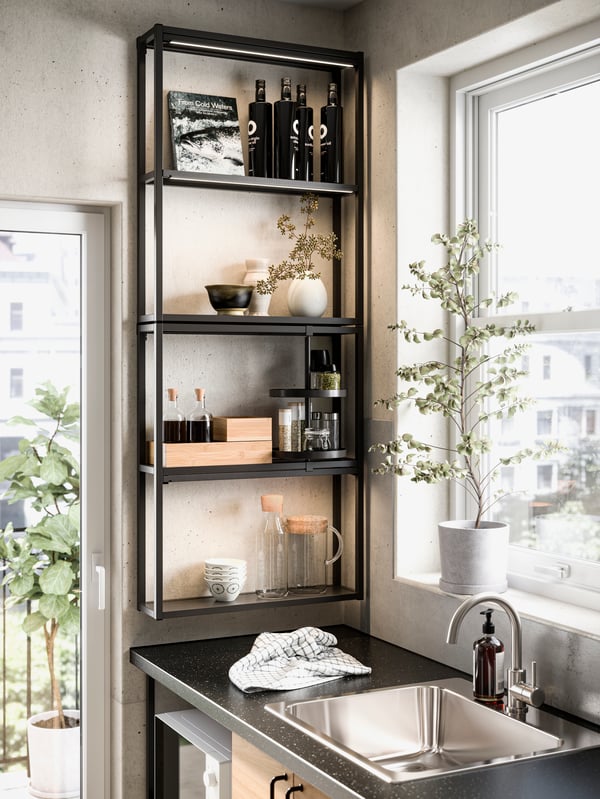 A kitchen corner with an ENHET wall frame with shelves in anthracite, by a window and sink, holding vases, boxes and bottles.