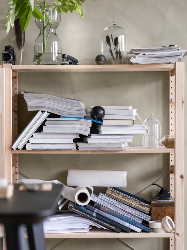 Wooden shelves against a wall with messy piles of books and magazines and glass vases with plants and feathers inside.