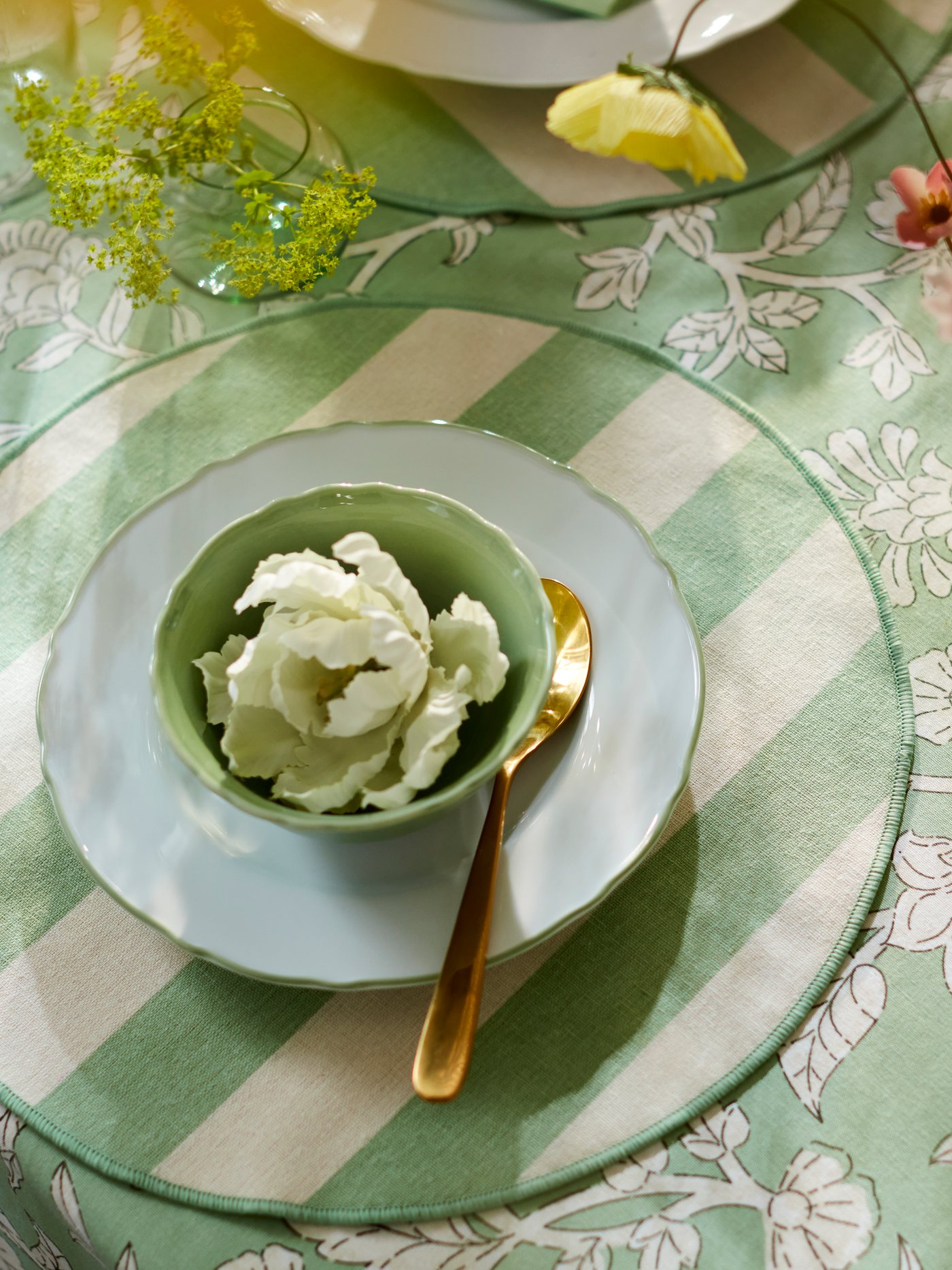 A green UPPLAGA bowl, decorated with a SMYCKA artificial spray, on a striped SMÖRFISK placemat and green SMÖRFISK tablecloth.
