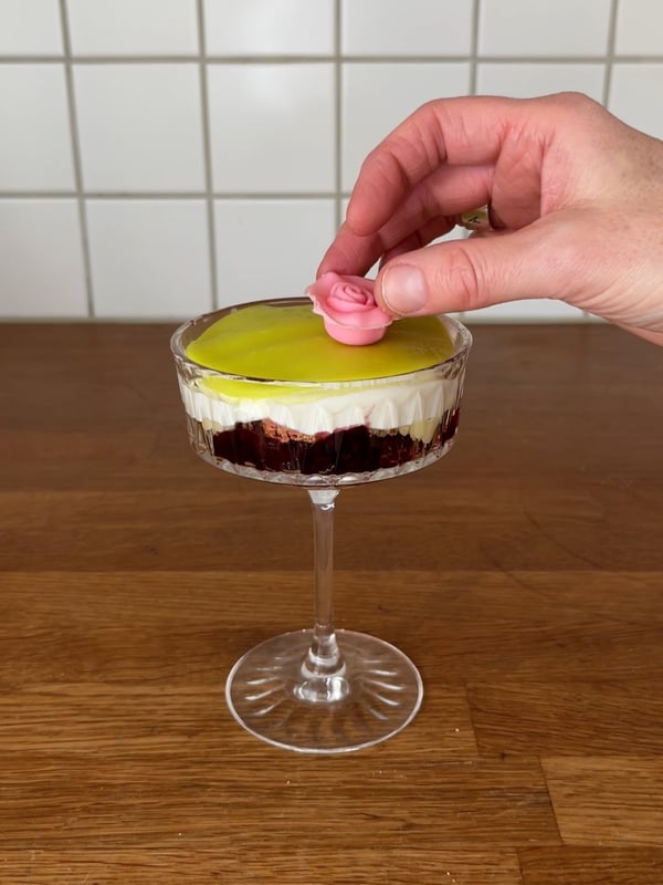 En champagnecoupe med tårtbotten, sylt, vaniljkräm och vispad grädde i. På toppen ligger ett marsipantäcke med en marsipanros på.