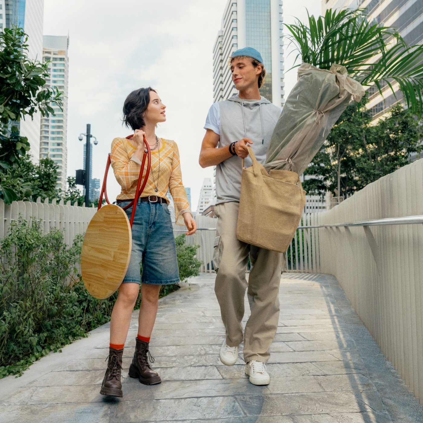 Two people are walking on a sidewalk carrying the KOMPISHÄNG side-table and plant pot. Behind them is a city skyline.