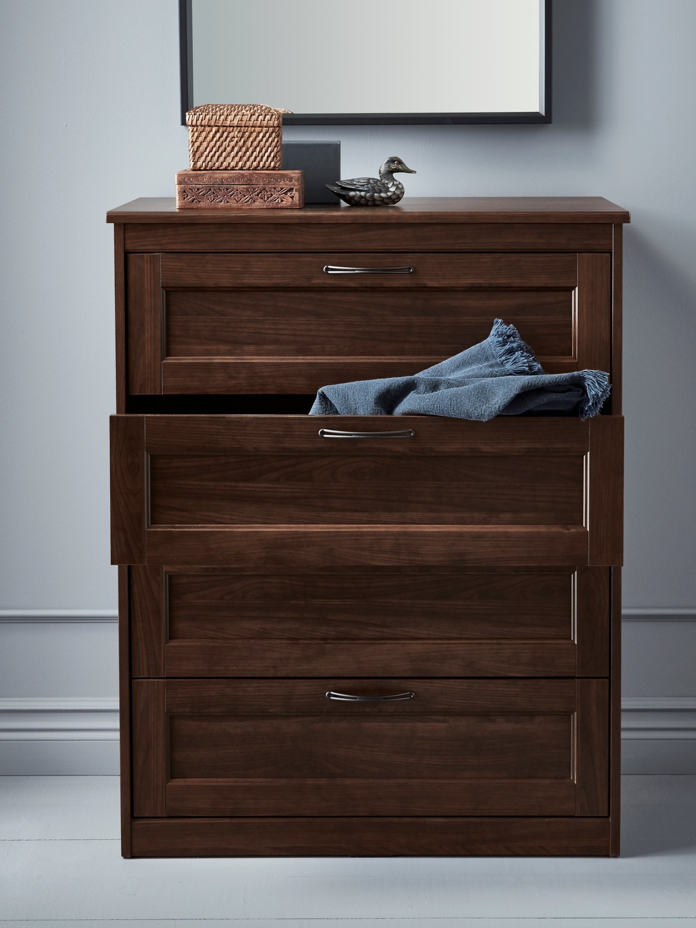 Bedroom with brown chest of drawers, boxes with lids, duck shaped decorative object, blue bedroom textile, mirror on wall.