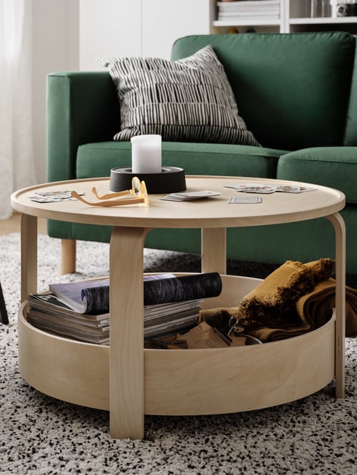 A round, light wood BORGEBY table in front of a green sofa with playing cards, a block candle and a pair of glasses on top.