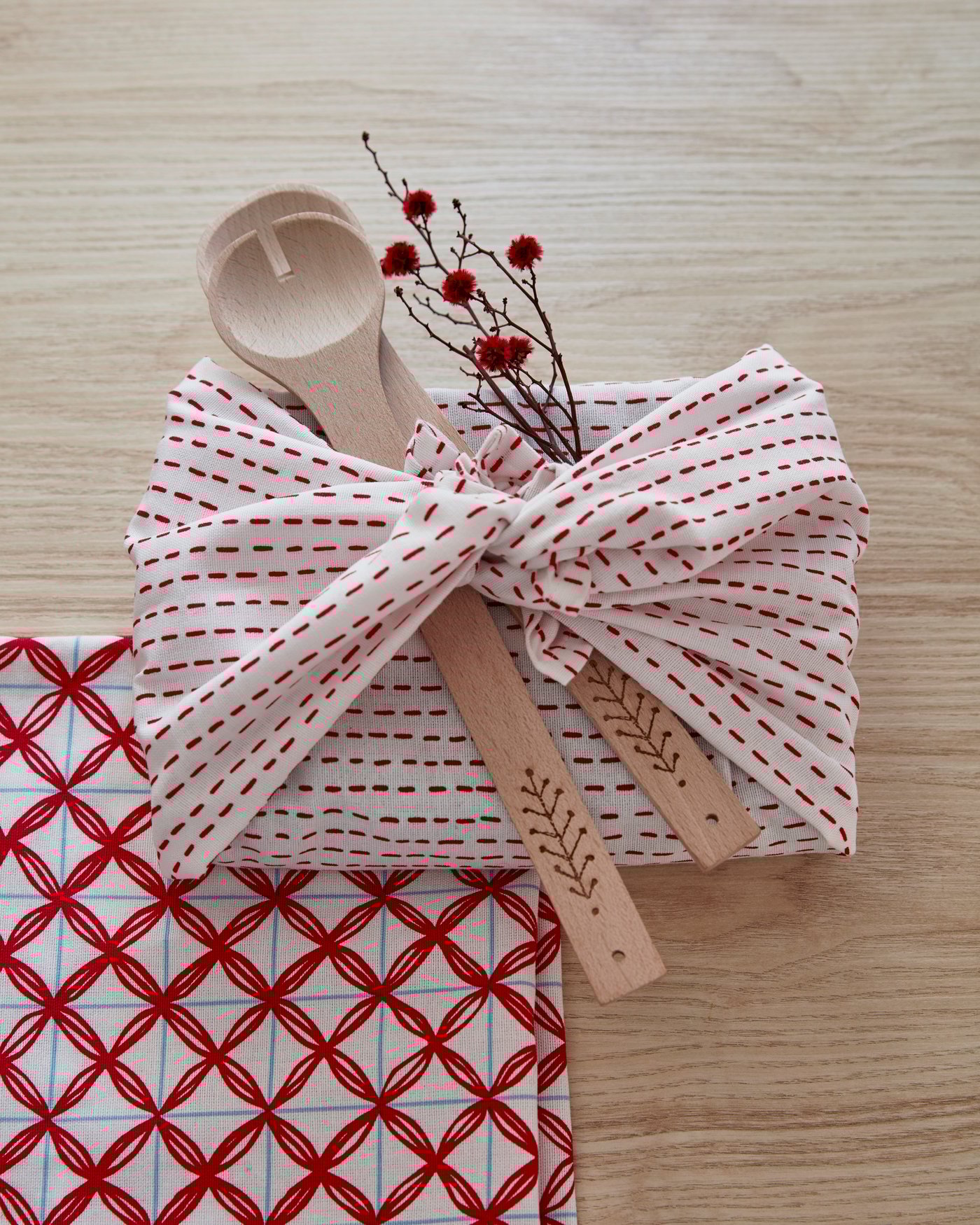 Ein rechteckiges Geschenk, verpackt in einem VINTERFEST Geschirrtuch und dekoriert mit Salatbesteck und einem Zweig.