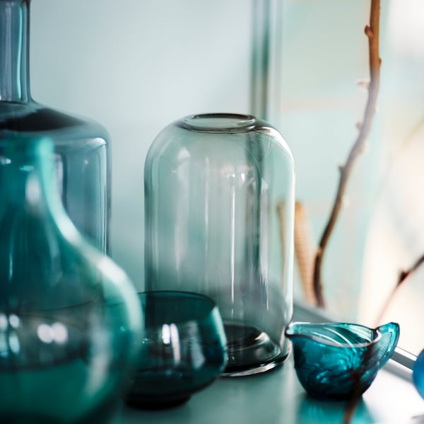 A grey DRÖMSK vase, other vases and other glass objects stand on a shelf in a RUDSTA glass-door cabinet.
