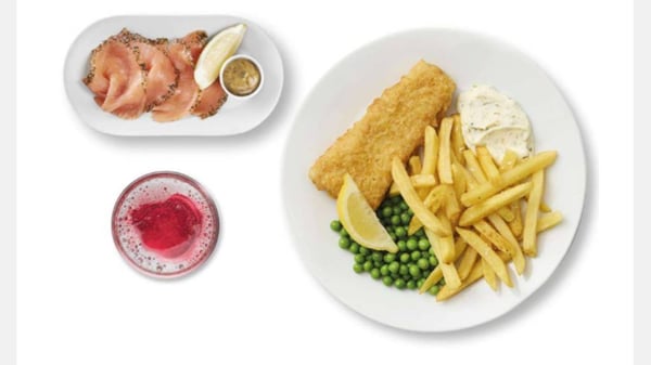 Assiette de poisson pané avec frites, petits pois, sauce et quartier de citron, accompagnée de saumon fumé en entrée et d’une boisson rouge.