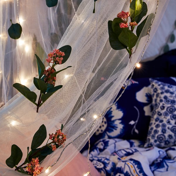 Delar av myggnät draperade över en madrass på en balkong där nätet är dekorerat med konstgjorda blommor och flera ljusslingor.