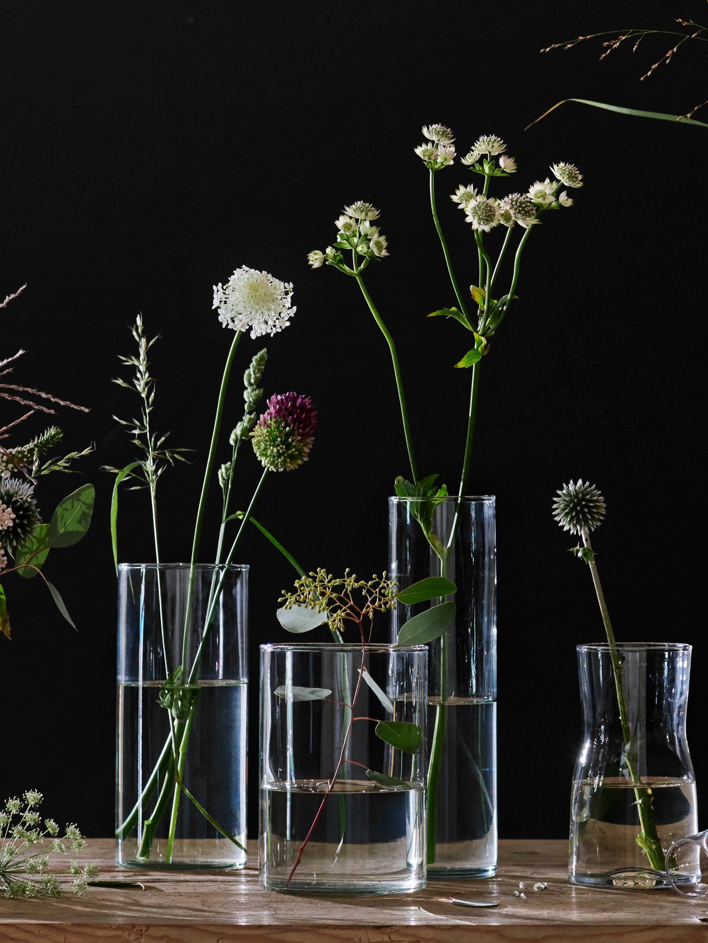 Pet staklenih vaza različitih visina s raznolikim cvijećem i biljkama, postavljenih na drvenoj površini ispred crne pozadine.