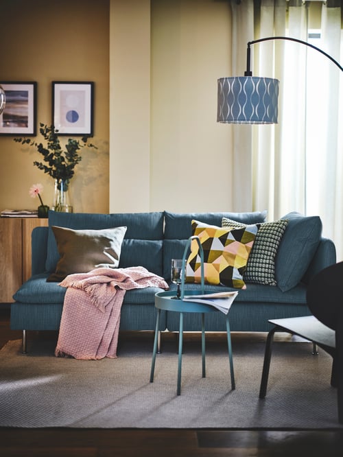 Salon z różową narzutą HORNMAL na niebieskiej sofie SÖDERHAMN. Sofa znajduje się obok okrągłego stolika i pod lampą podłogową.