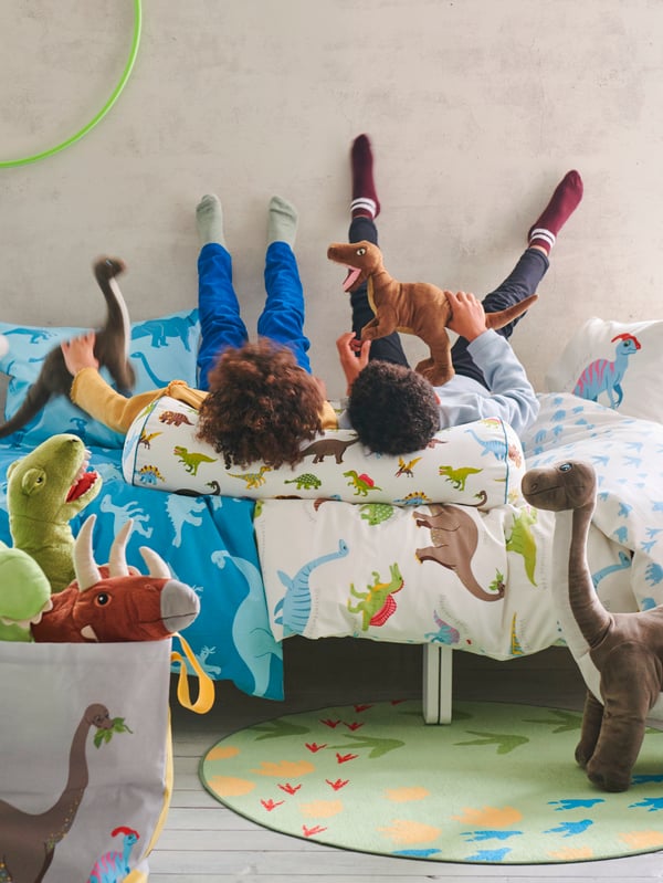 Kinderzimmer mit zwei Kindern die auf buntem Dinosaurier-Bett spielen umgeben von vielen Stofftieren