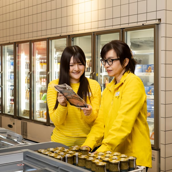 フードマーケットの冷凍ケース前で、黄色いユニフォームのイケアのコワーカー2人が商品パッケージを確認している
