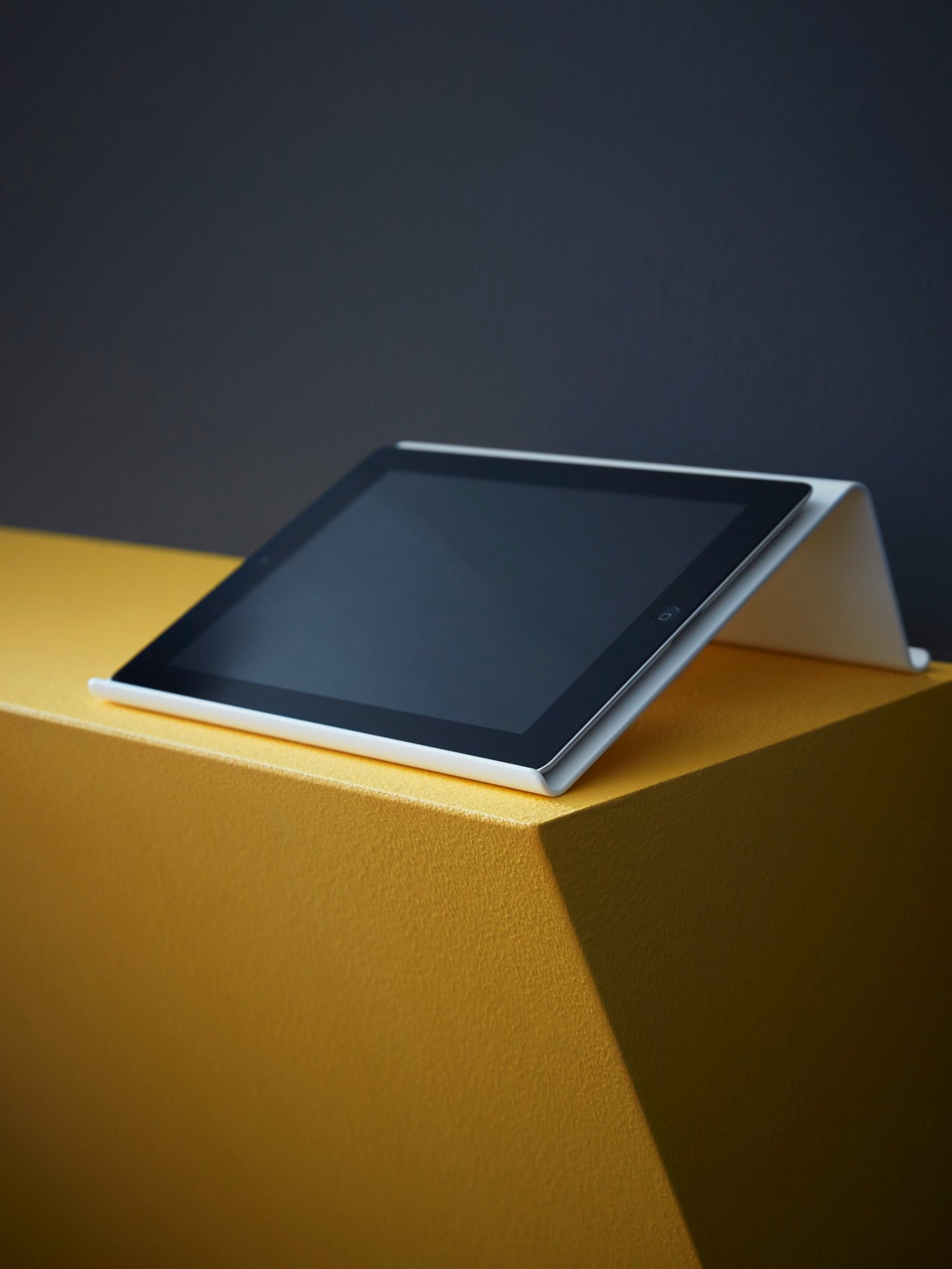 A tablet lies on a white ISBERGET tablet stand which stands on a low wall that has been painted dark yellow.