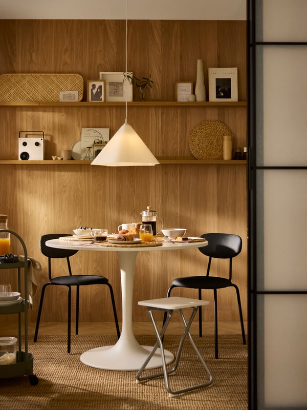 Drawn-apart SKYTTA sliding doors open onto a wood-wall dining area centred around a white DOCKSTA table set for breakfast.