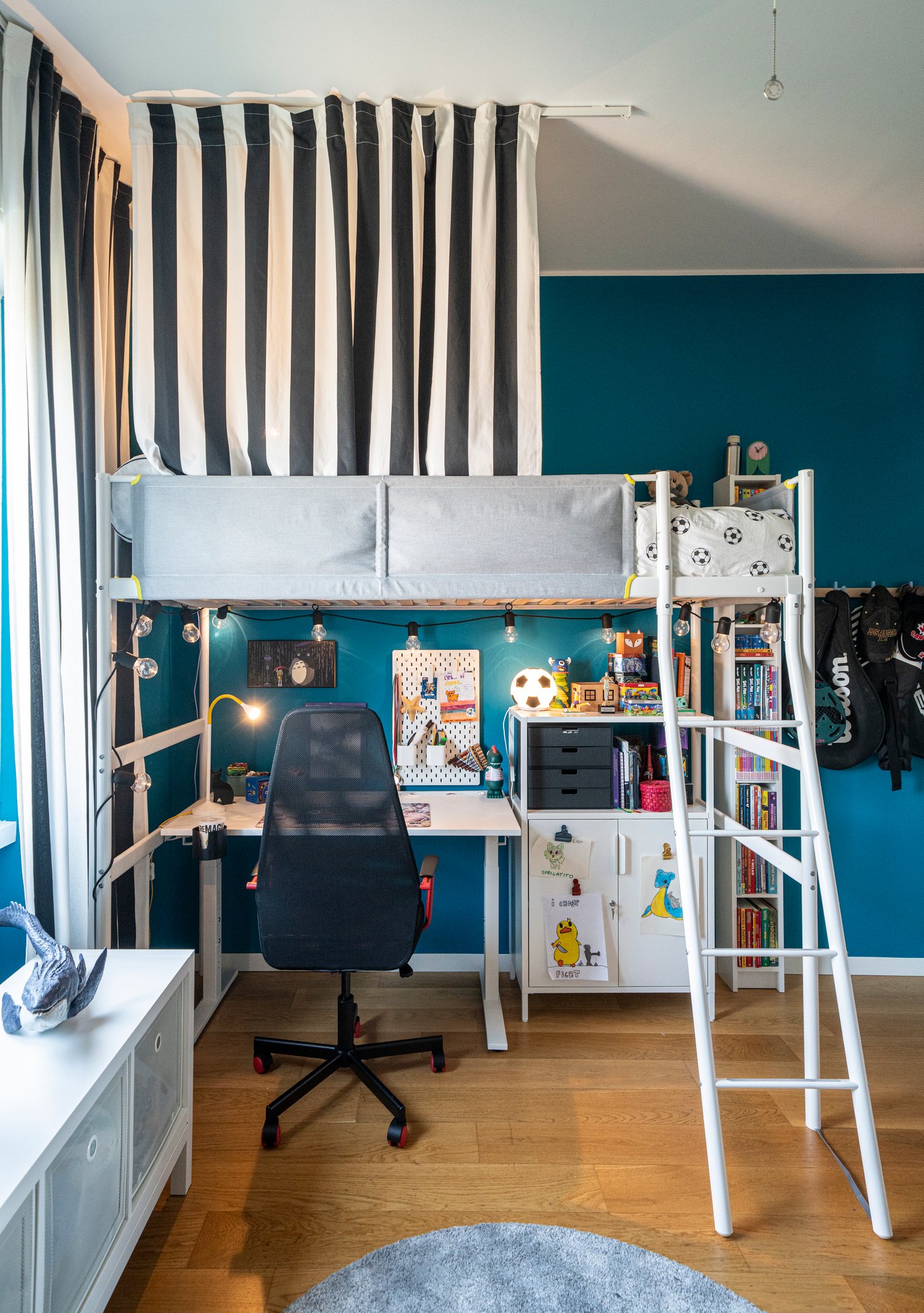 Cameretta con letto a soppalco bianco e tenda a righe, scrivania con sedia da studio sotto il letto, librerie piene di giochi e un gatto nero sdraiato sul tappeto rotondo.