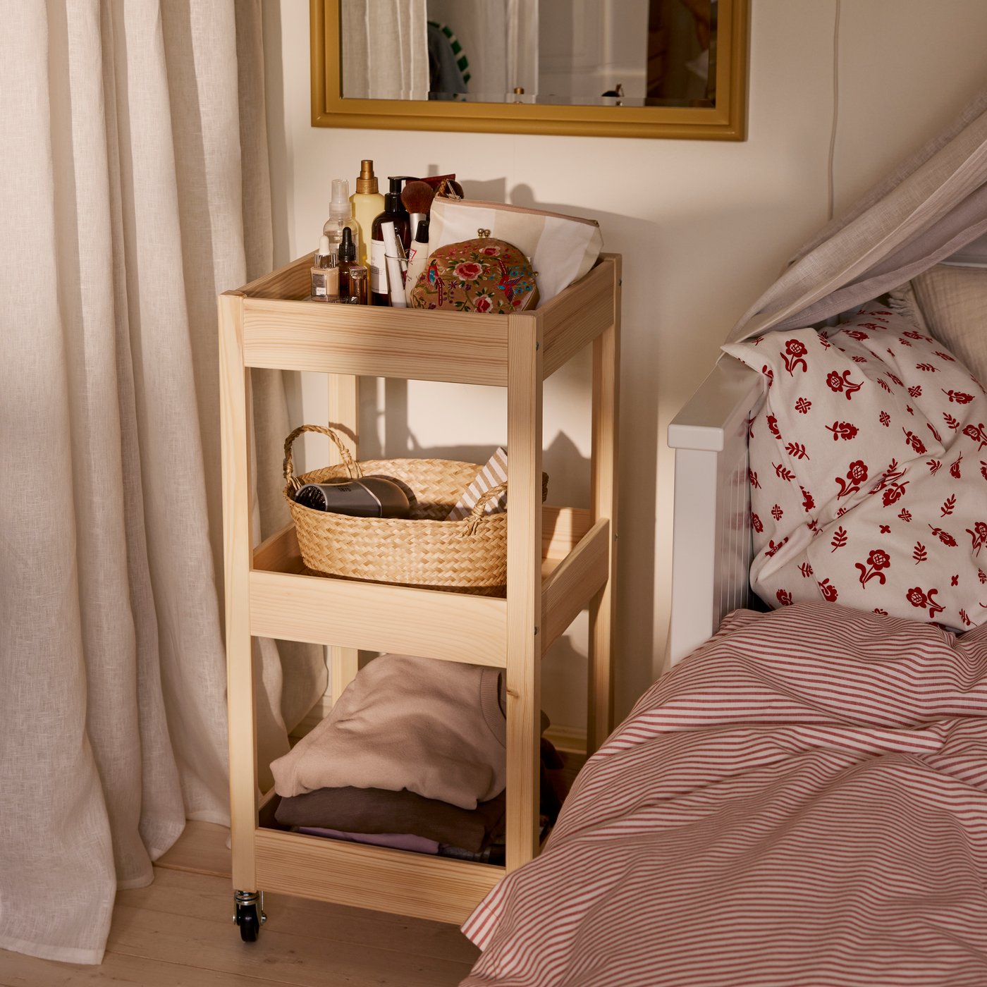 Close-up of LOSHULT trolley in pine next to a bed. A mirror above it, and the trolley is full of accessories and things.