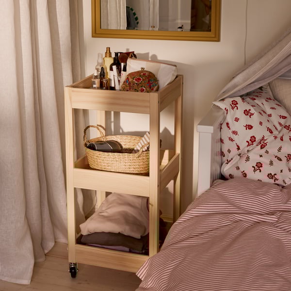 Close-up of LOSHULT trolley in pine next to a bed. A mirror above it, and the trolley is full of accessories and things.