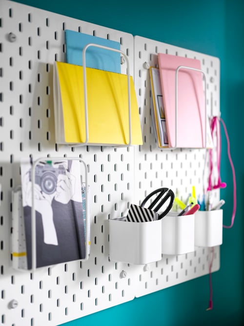 A teal wall has two, side-by-side white SKÅDIS pegboards with notebooks in letter holders and pens in containers.