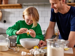 Chłopak rozbijający jajko do białej miski w towarzystwie mężczyzny stojącego obok niego.
