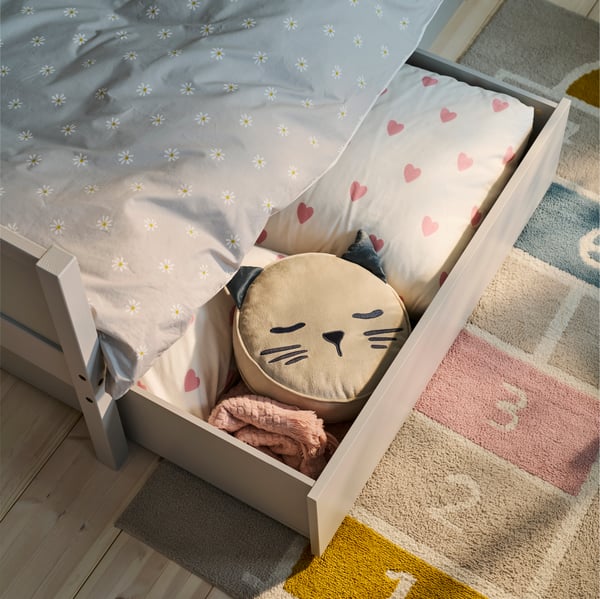 SMYGA-Bett mit Aufbewahrungsschublade, BARNDRÖM-Bettwäsche mit Herzmuster und Katzenkissen, bunter Hüpfspielteppich daneben.