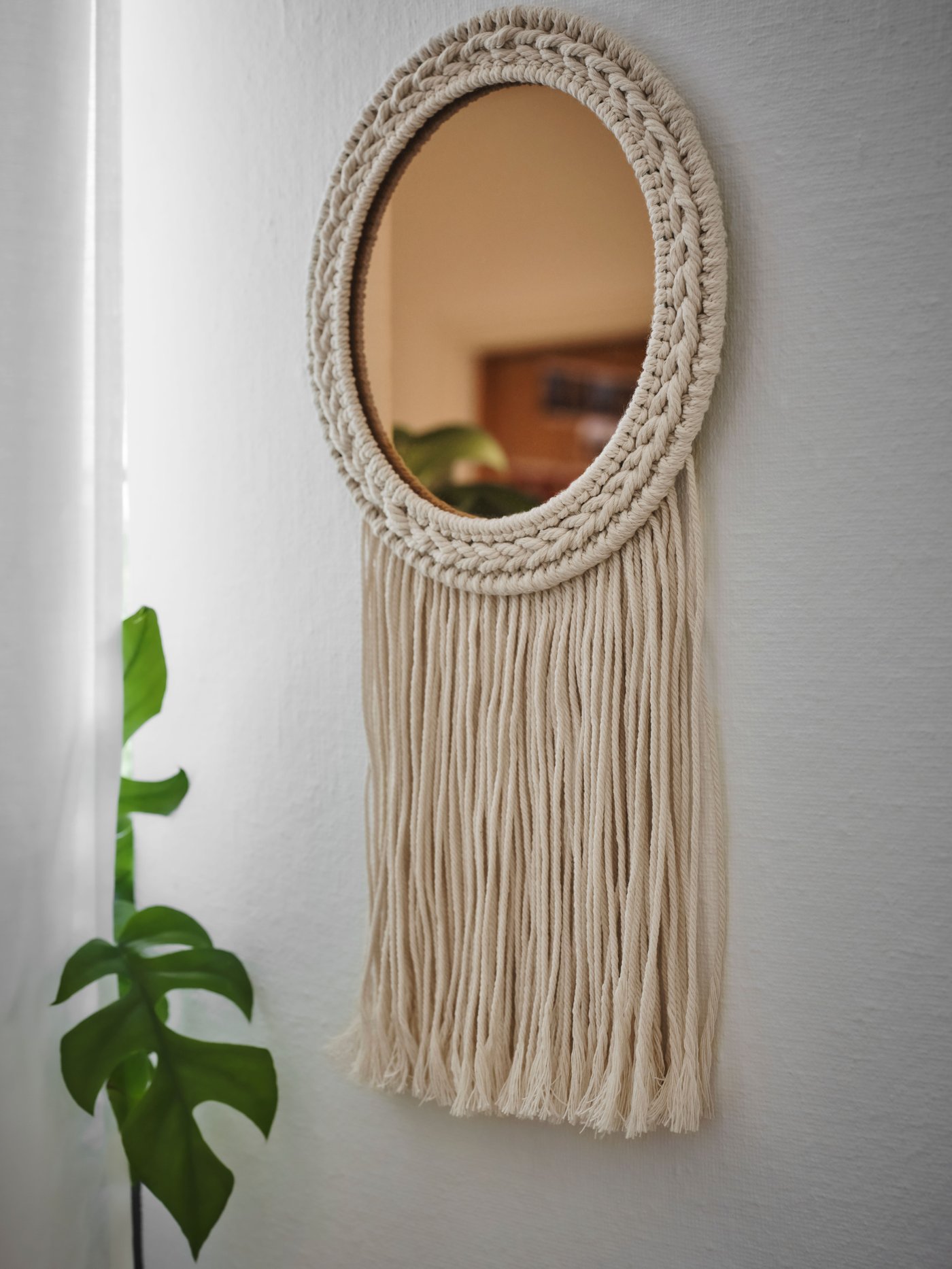 ENERGISKOG okrasno ogledalo v bež barvi in barvi bakra z resicami visi na steni blizu velike rastline in zavese.