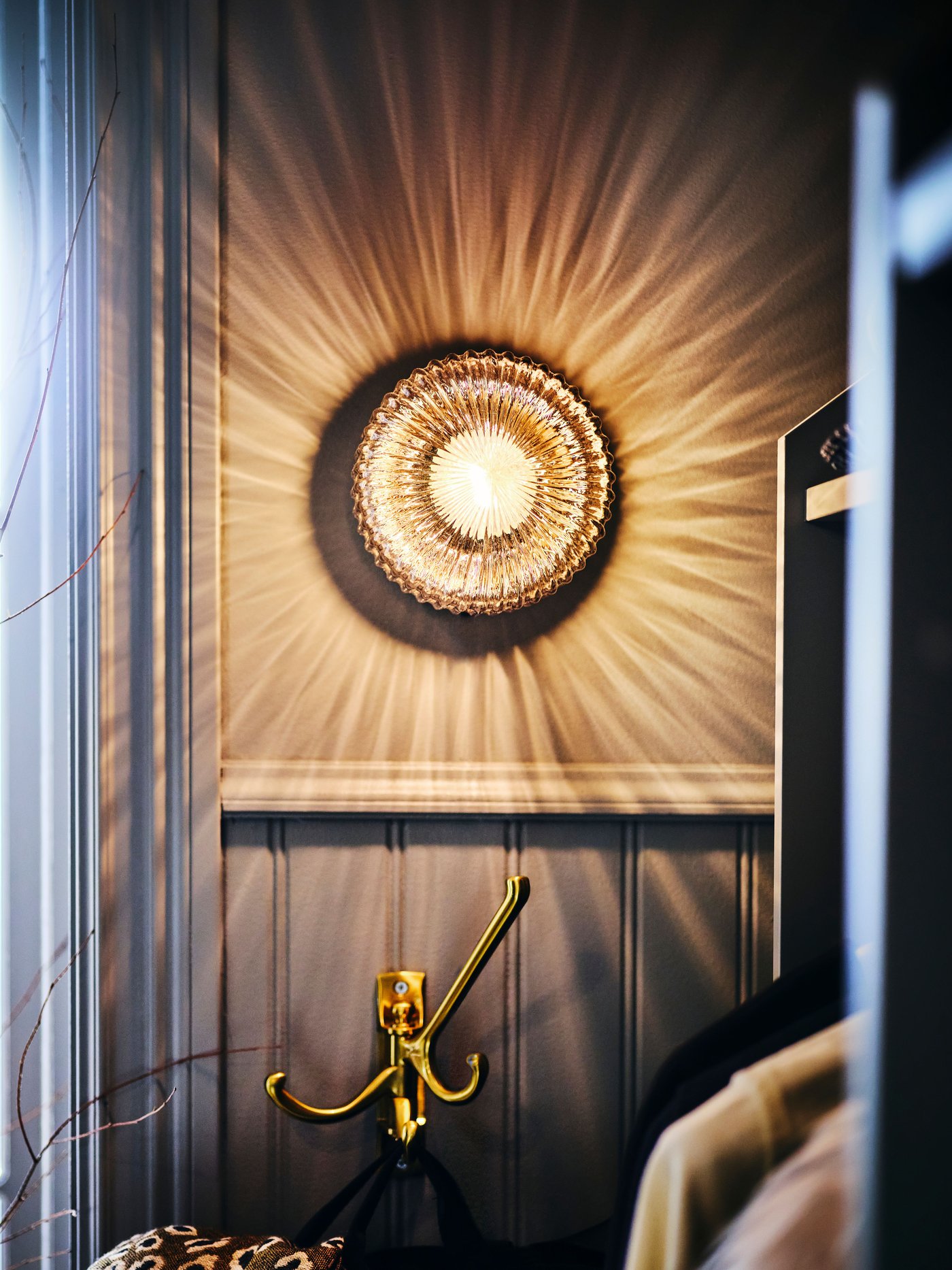 A lit SOLKLINT wall lamp with a brass KÄMPIG 3-arm swivel hook on the wall below, with clothes hanging to the right.