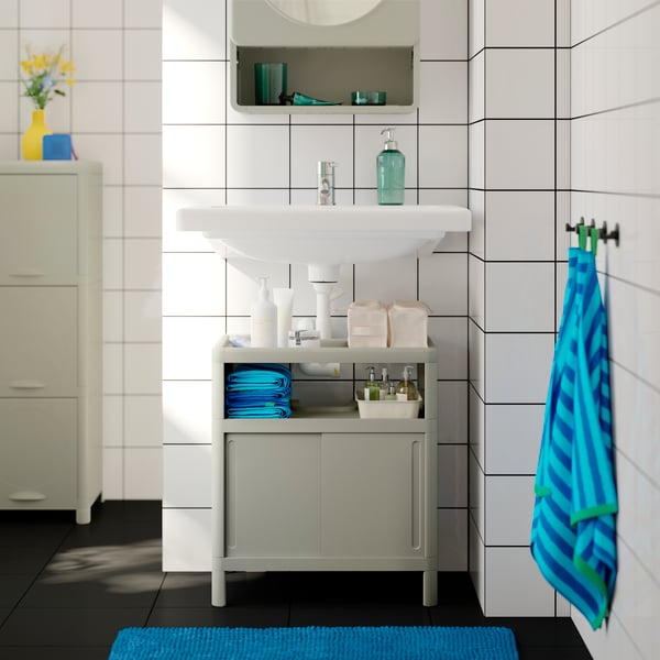 A green IVÖSJÖN wash-basin cabinet with sliding doors is under a sink. A blue mat is by and striped towels are near the sink.